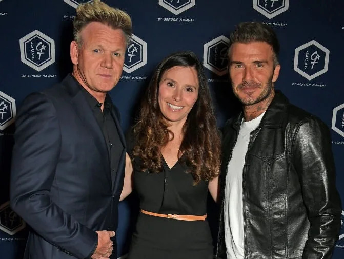 Three people pose on a red carpet: one in a suit, one in a dress, and one in a leather jacket with jeans, smiling for the camera