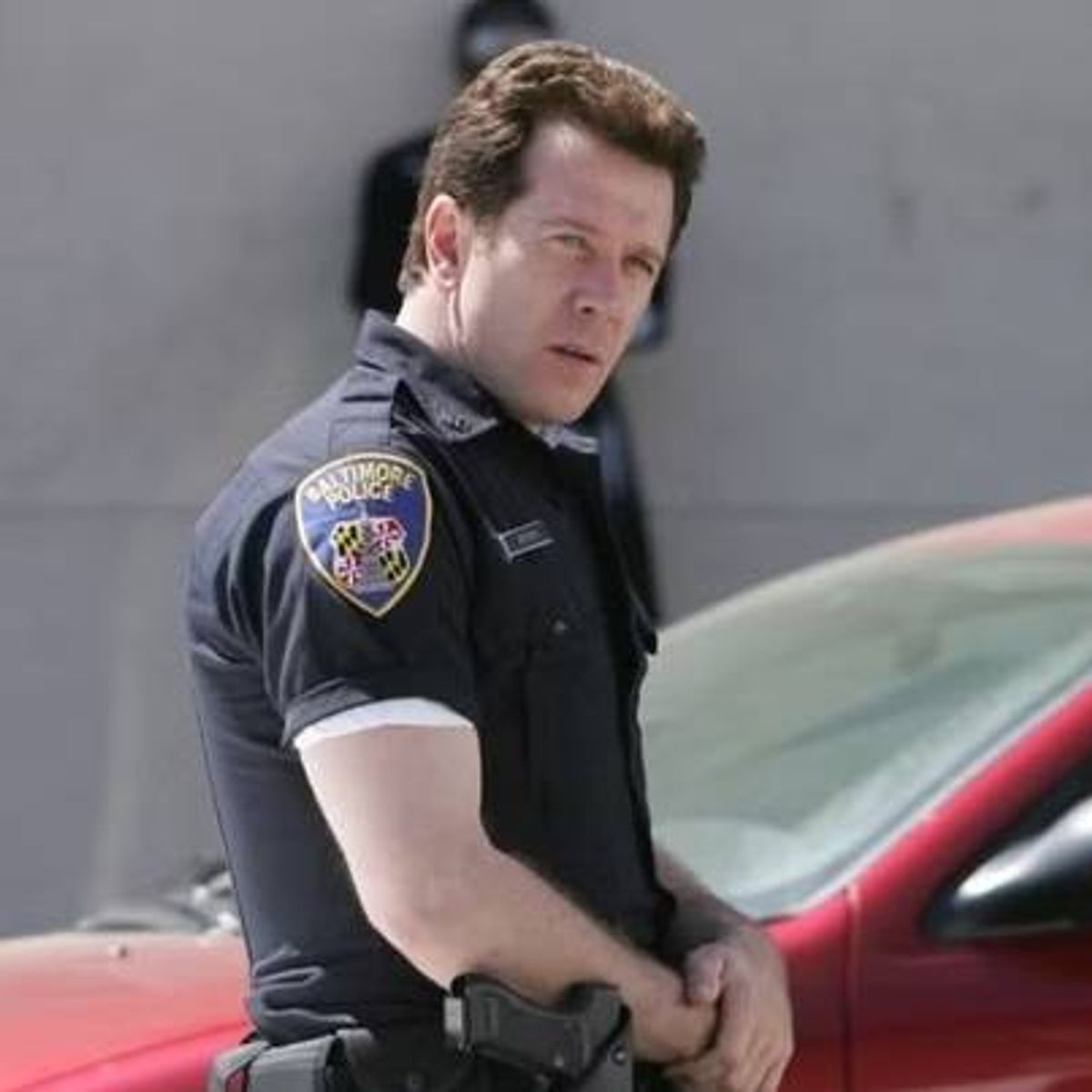 An individual in a police uniform stands outside near a vehicle, with a serious expression on their face, and a building in the background.