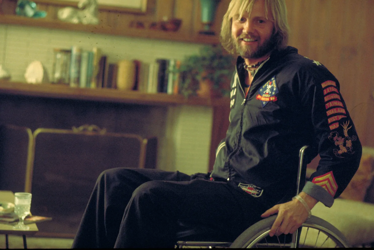 A man with blond hair and a beard, wearing a black jacket with colorful patches, sits smiling in a wheelchair in a cozy living room with bookshelves and wood-paneled walls in the background.