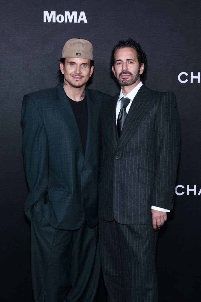 Marc Jacobs wears a traditional black pinstripe suit standing next to partner Char Defrancesco in a black jacket, black t-shirt and hat.