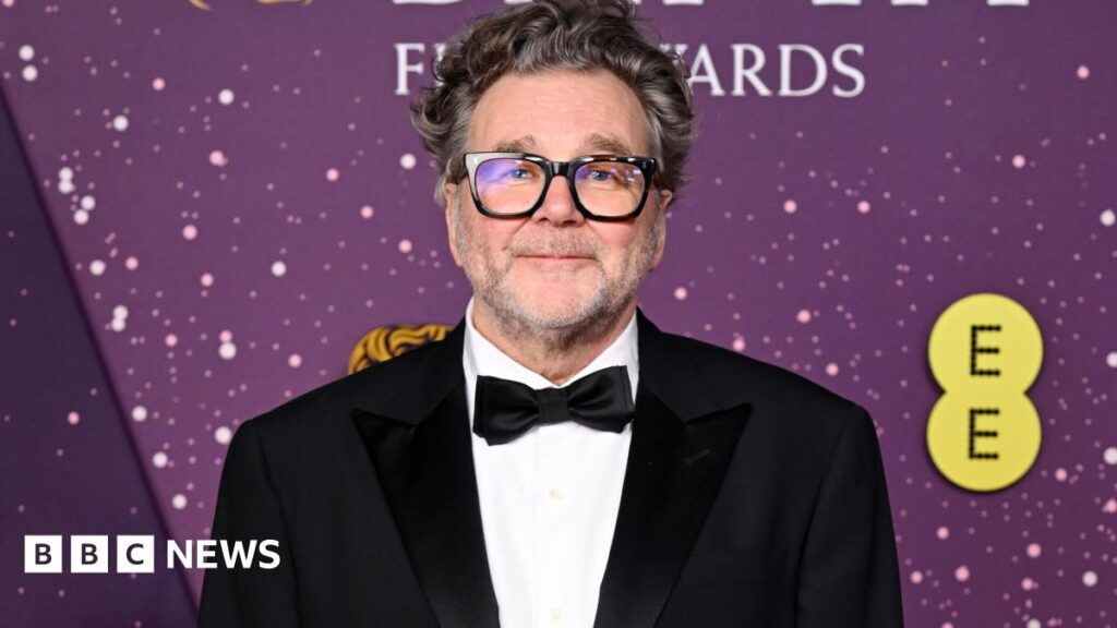 Kirk Jones. He has short grey hair and stubble. He is wearing black rimmed glasses, a white shirt, a black suit jacket and a black bowtie. He is looking directly at the camera and smiling.
