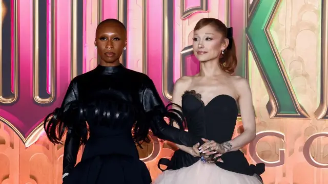 Cynthia and Ariana in fancy outfits standing holding hands. Cynthia looks in front of her in a serious manner, Ariana looks tot he side with a small smile