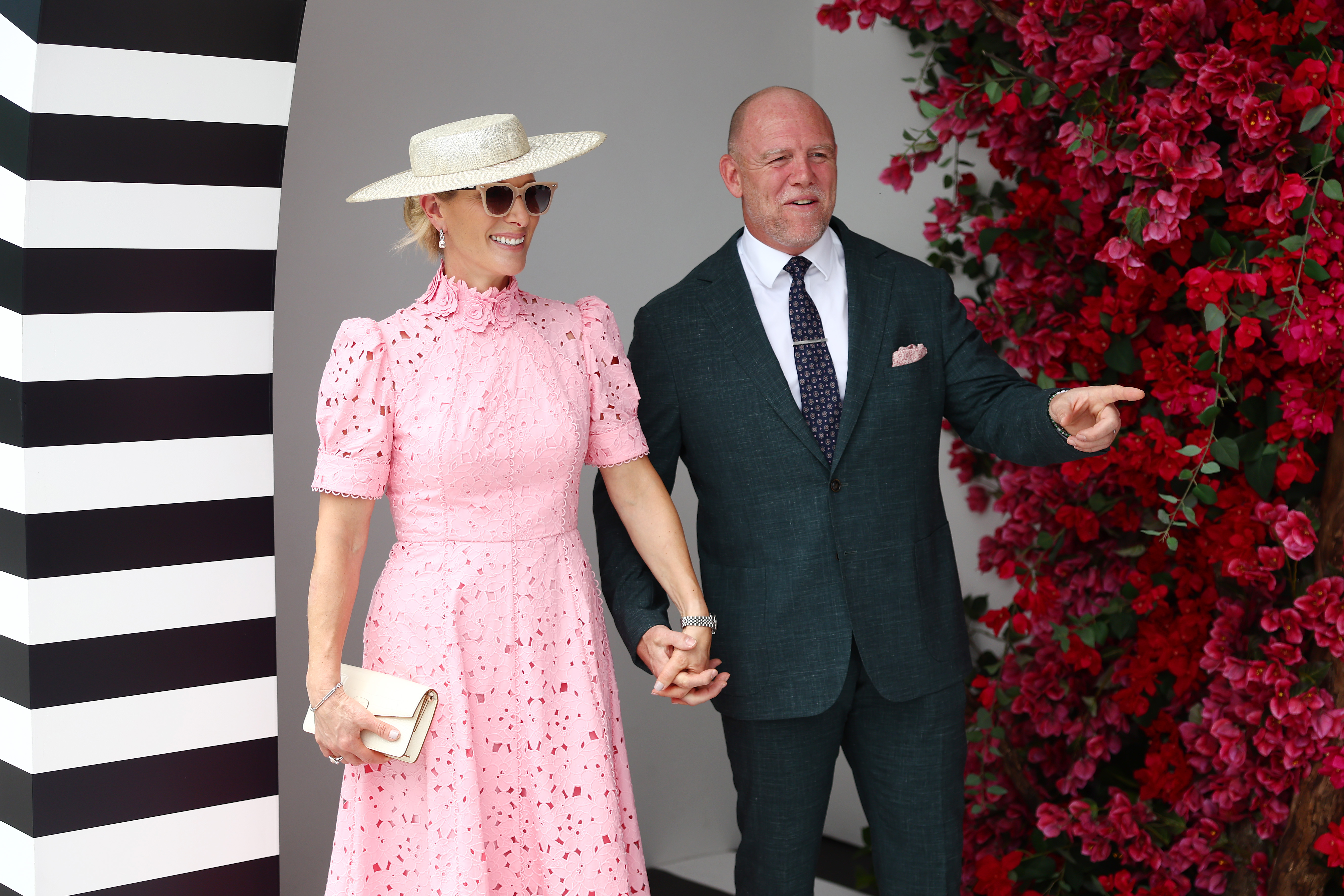 Zara Tindall wearing a pink dress and holding hands with Mike Tindall next to a wall of flowers