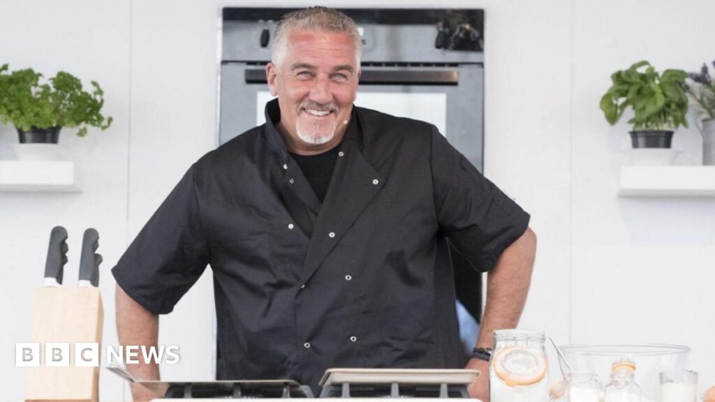 Paul Hollywood: The Wirral-born baker who rose to fame on GBBO Paul Hollywood has short grey hair and is wearing a black t-shirt and a black chef's coat. He is stood in a kitchen has his hands on a work surface. A set of knifes and glass containers are in the foregrounds and an oven and two potted plants are in the background.
