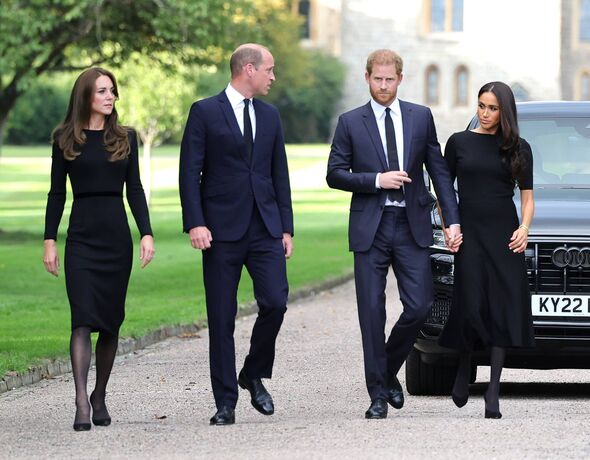 Four individuals, dressed in formal attire, are walking together on a pathway. Two of them are men, both wearing suits and ties,