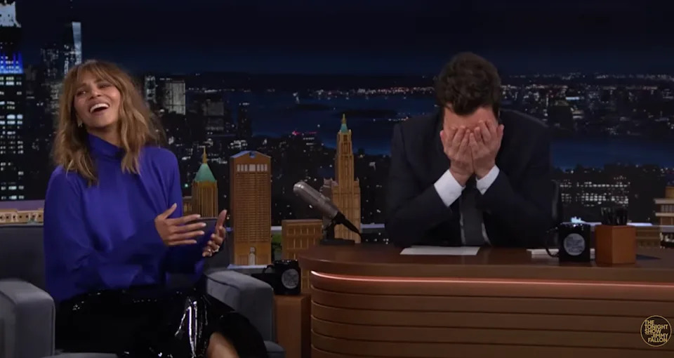Halle Berry in a blouse and skirt laughs, while Jimmy Fallon in a suit covers their face with their hands on The Tonight Show set
