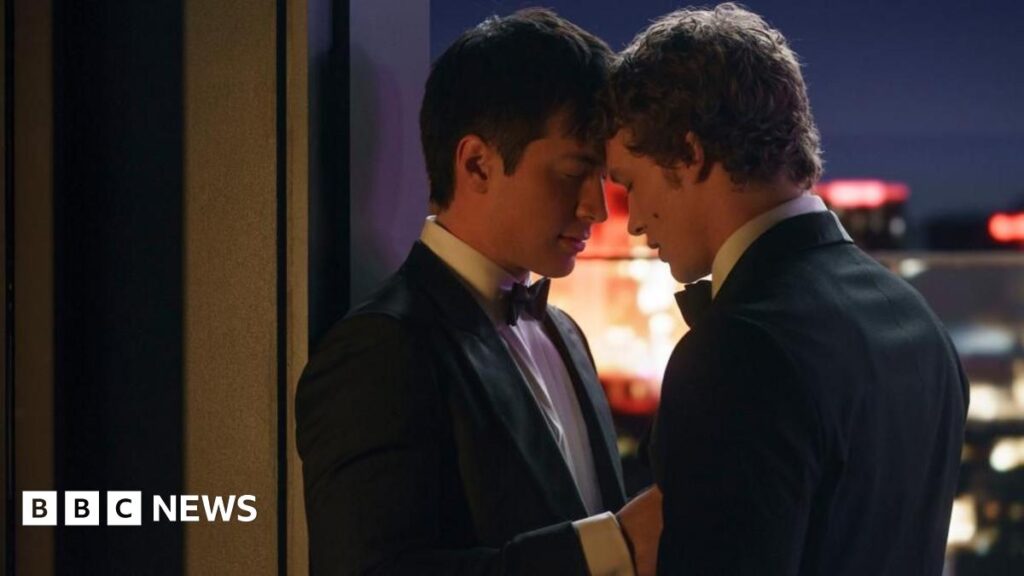 Prime Minister Mark Carney, wearing a beige fleece sweater with a red Canadian maple leaf on the arm, laughs as he hugs Heated Rivalry actor Hudson Williams, wearing a leather jacket.