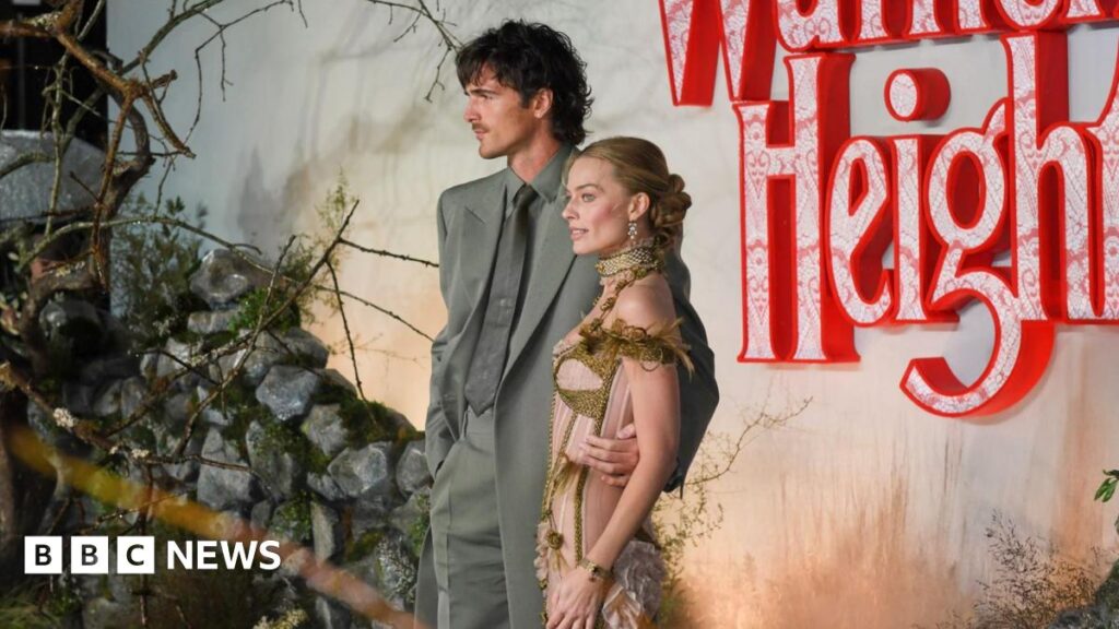 Margot Robbie and Jacob Elordi pose before a moody “Wuthering Heights” backdrop of red lace‑patterned letters, rocks, and branches; one wears a grey suit, the other an ornate gold‑and‑green outfit.