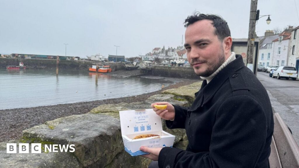 ‘I based horror video game on working in a Scottish chippy’ James Muirhead, who is wearing a dark coat, sits by a harbour on a grey day, holding a takeaway box of fish and chips and a lemon wedge. Fishing boats rest in the background near a stone pier, with pastel-coloured houses lining the waterfront.