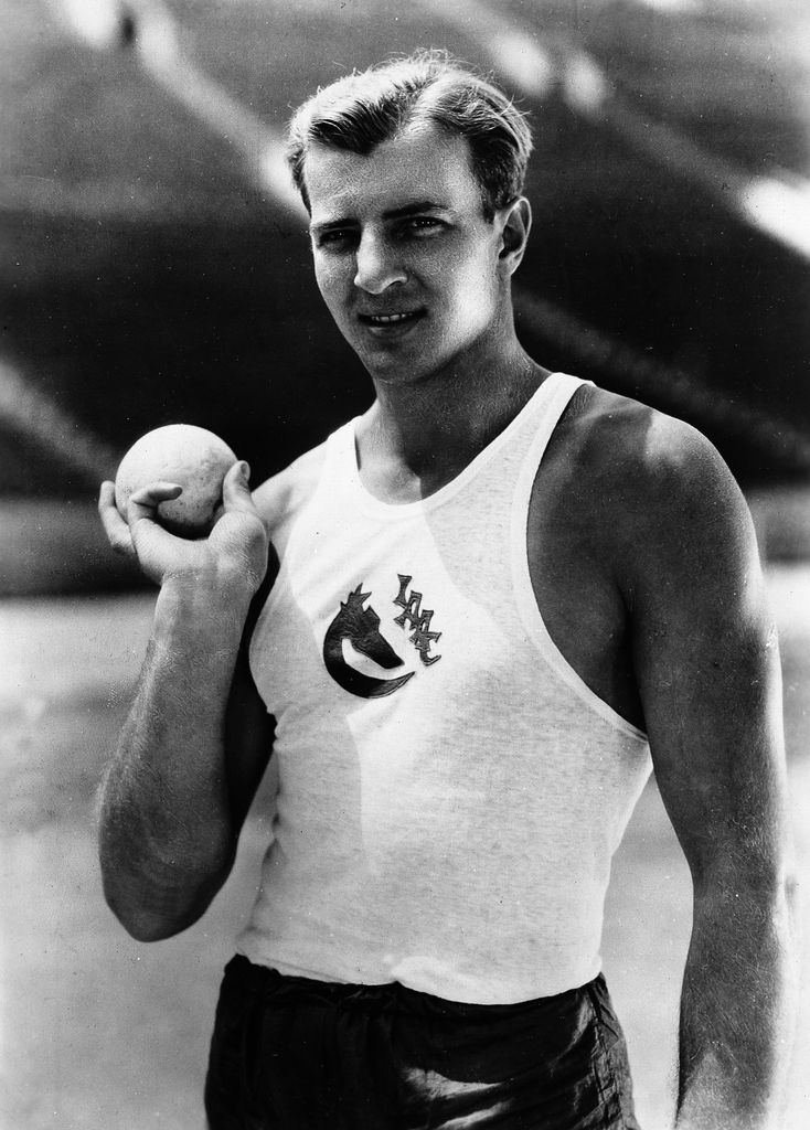 Shot-putter and actor Bruce Bennett holds a ball in one hand held back against his chest.