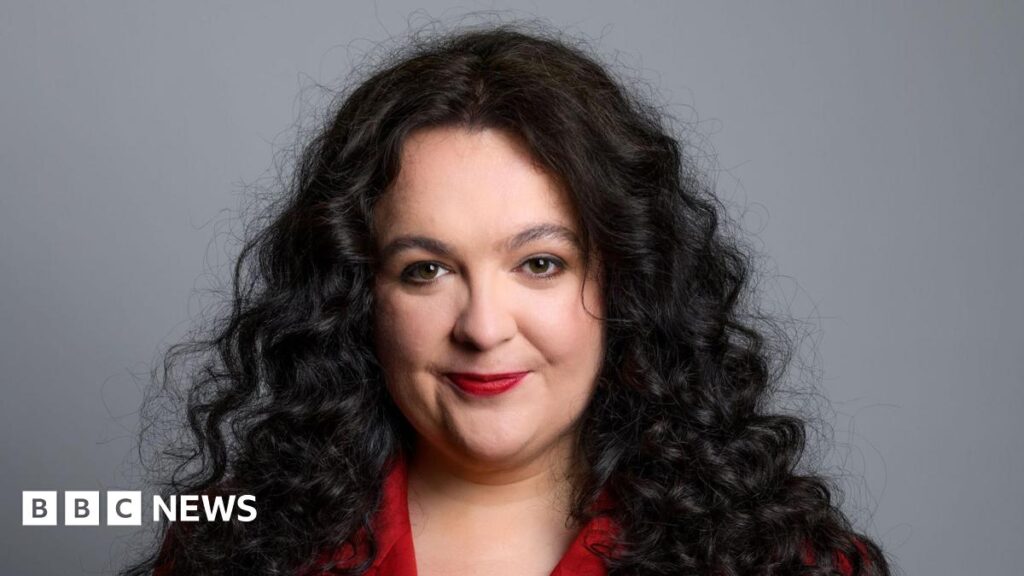 Ashley Storrie, a woman with long dark hair, wearing a red jacket. She is staring into the camera