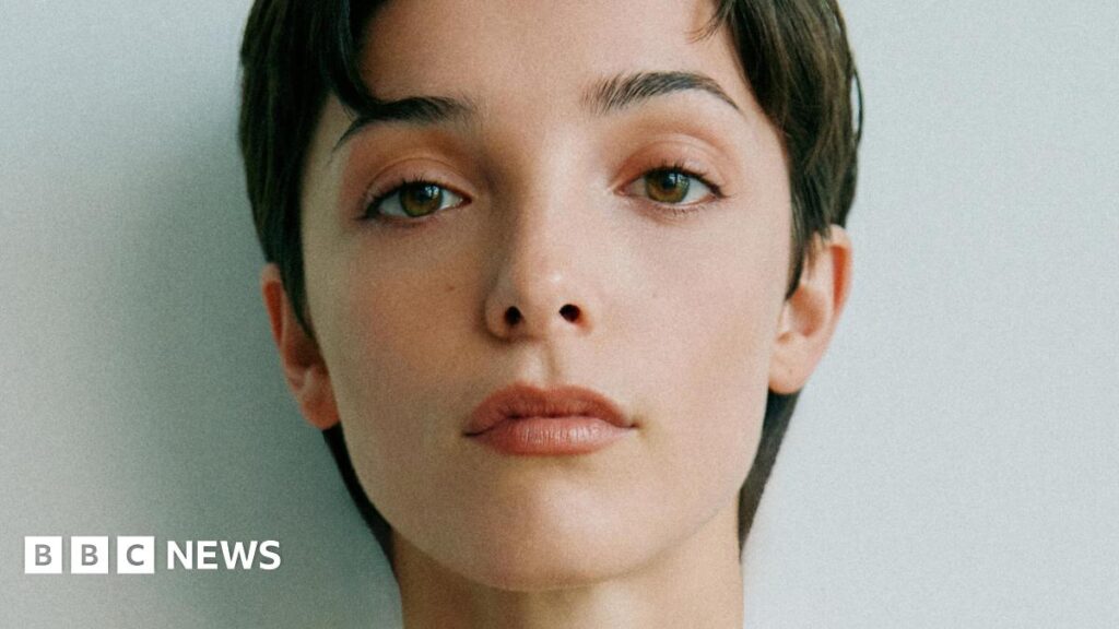 Jessica Reynolds, a woman with short, dark hair, looks to the camera with a neutral expression for a close-up photo.  She has brown eyes and is lightly made up.