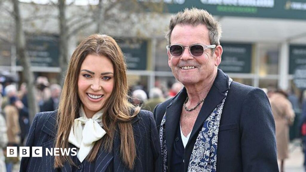Lorna Luxe: Stars pay tribute to her husband John Andrew’s death A woman with long-brown hair and wearing a dark pinstriped suit with a white scarf stands next to a man with a dark coloured suit and blue and white patterned scarf. He has short grey hair which is swept back and spiked up. He has a chain necklace and sunglasses. They are both smiling.