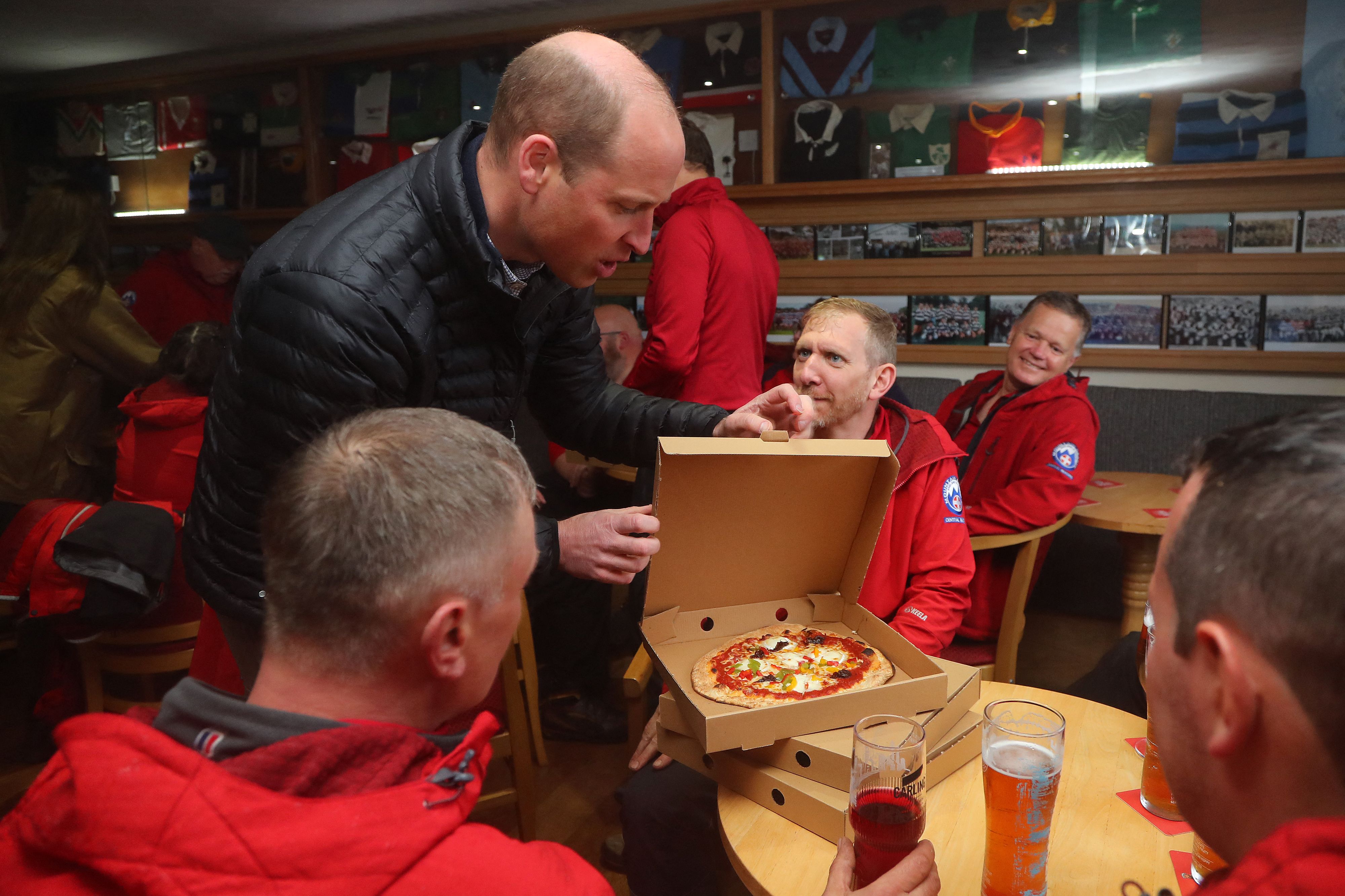 Prince William opening a pizza box surrounded by men sitting at tables