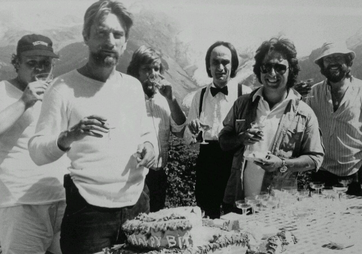 Six men stand outdoors by a table with a birthday cake and drinks, mountains in the background. Some smile or eat, one wears a bow tie, others wear casual clothes and sunglasses. The setting appears to be a celebration.