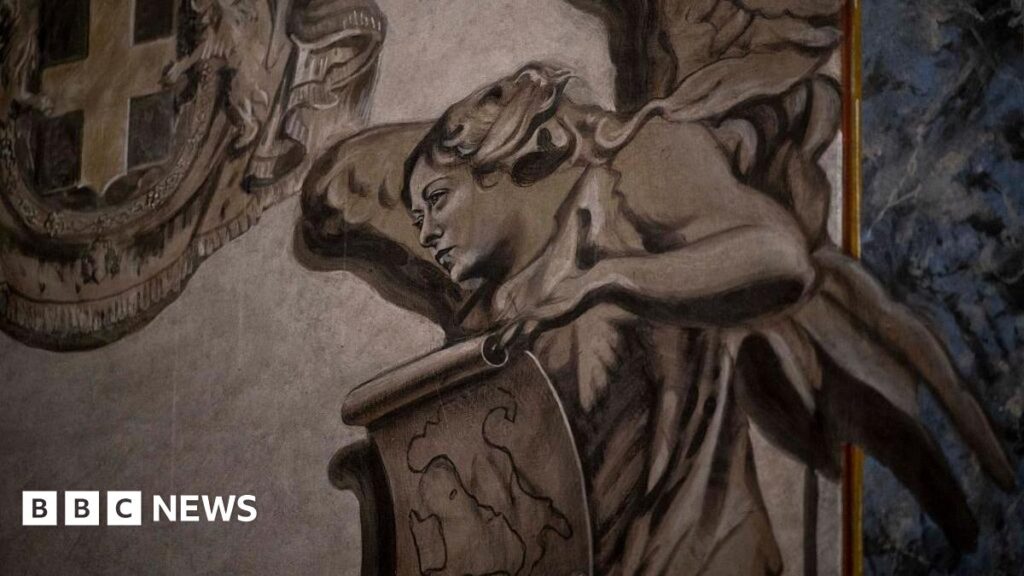 Restored angel fresco resembling Italian PM Meloni sparks investigation A close-up shot of a painted grey angel. Its face resembles that of Italian Prime Minister Giorgia Meloni. She has wings and holds a scroll on which the shape of Italy is drawn.