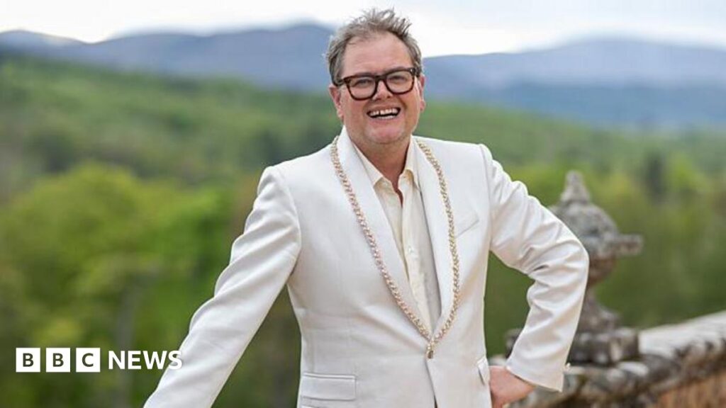 Why has Alan Carr bought a castle and where is it? Alan Carr, a man with unkempt hair and thick-rimmed glasses. He is standing with one hand on his hip and the other leaning on a wall in front of lots of greenery. He is smiling which shows off a gap in his teeth. He is wearing a cream suit jacket with gold beading on it.