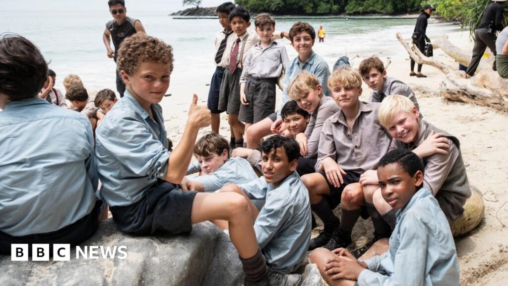 A group of young boys are sat down on a beach on a remote island. They are posing at the camera. One boy is sitting on a rock, putting his thumb up at the camera.