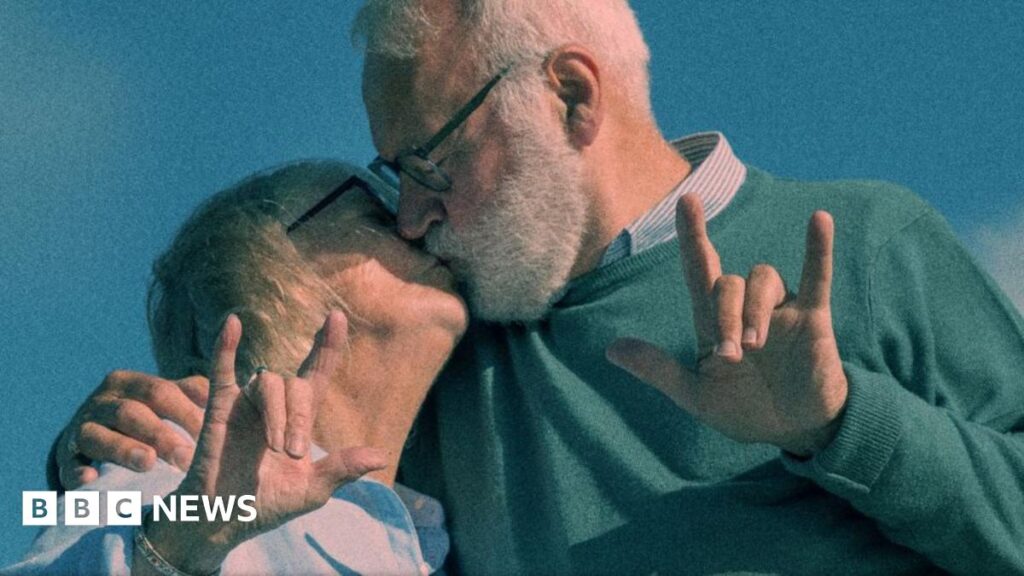 Two young baristas, wearing black aprons with the Dialogue cafe logo on the front - which includes text in white and a large purple heart - stand in front of the café's windows.
