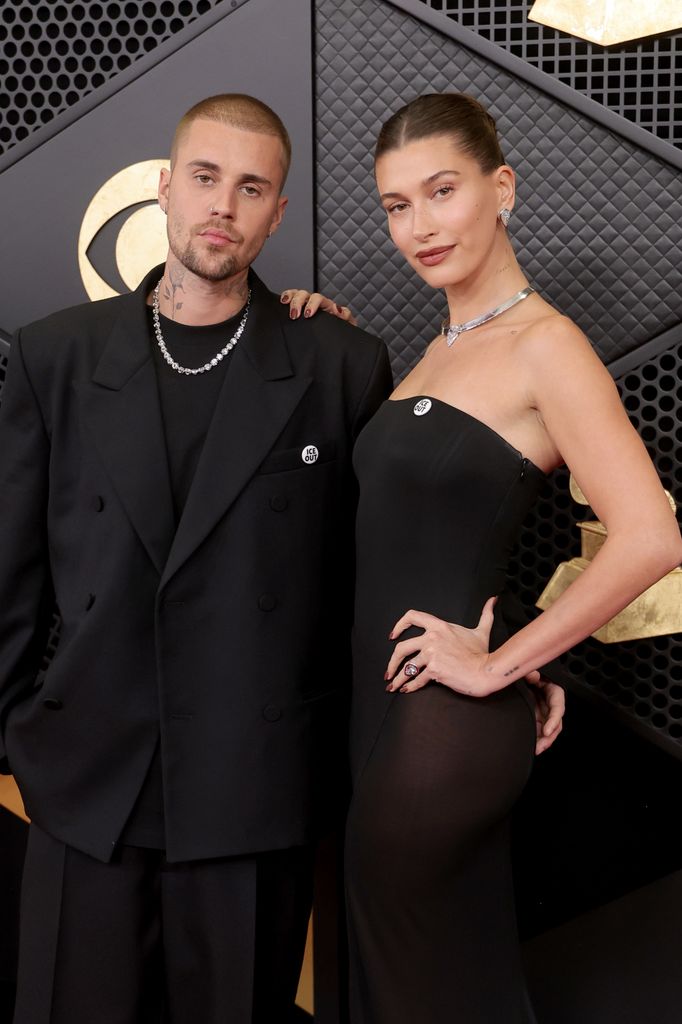Justin Bieber wears a black suit with a silver chain and a buzzcut. He stands next to wife Hailey Bieber who wears a strapless black dress and a silver necklace with one hand on her hip and the other on his left shoulder.
