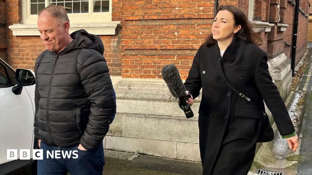 Raymond McDonald, a man in his fifties, is walking away from Philippa Goymer who is a BBC journalist. She is dressed in black and has long dark hair and is carrying a large microphone. He has a black jacket, blue jeans and has his hands in his pockets. Behind them both is a brick built building and a white car is parked to the side.