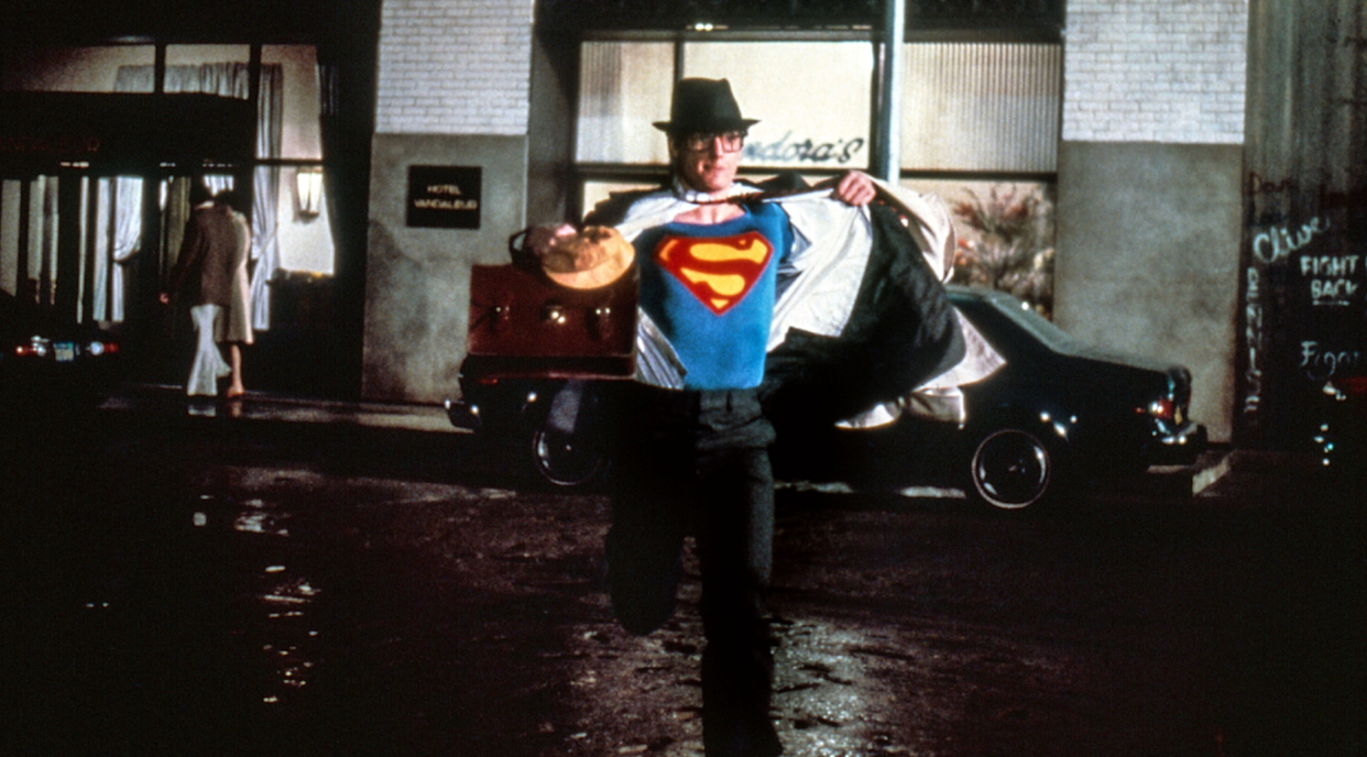 A man in a suit and hat runs down a city street at night, pulling open his shirt to reveal a Superman costume underneath.