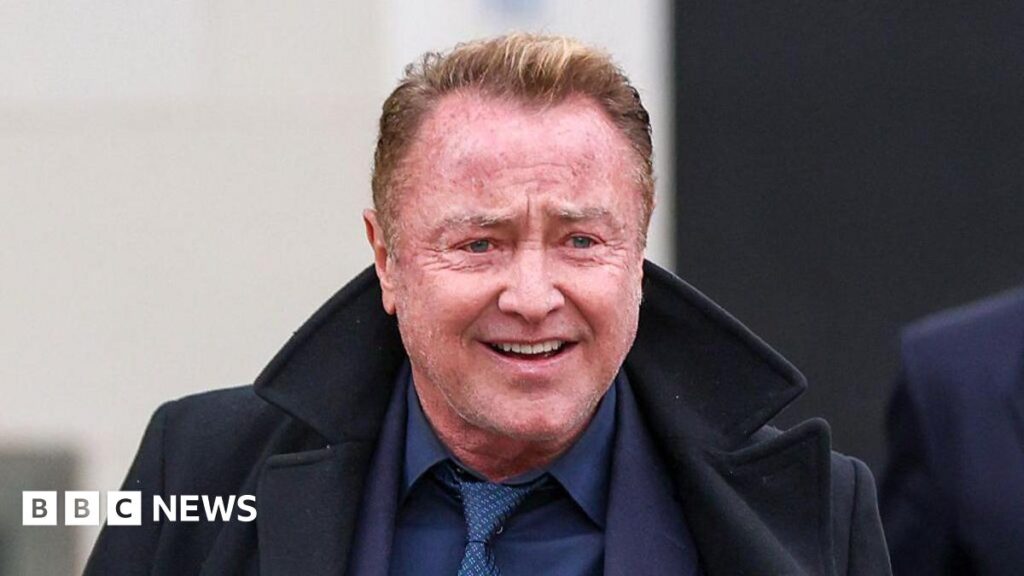 Michael Flatley. He has short reddish hair, wearing a navy suit and black coat.