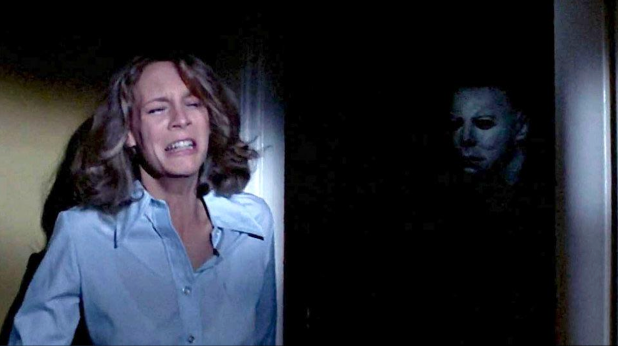 A frightened woman in a blue shirt stands in a doorway, while a masked figure lurks menacingly in the dark background, partially obscured by shadows.