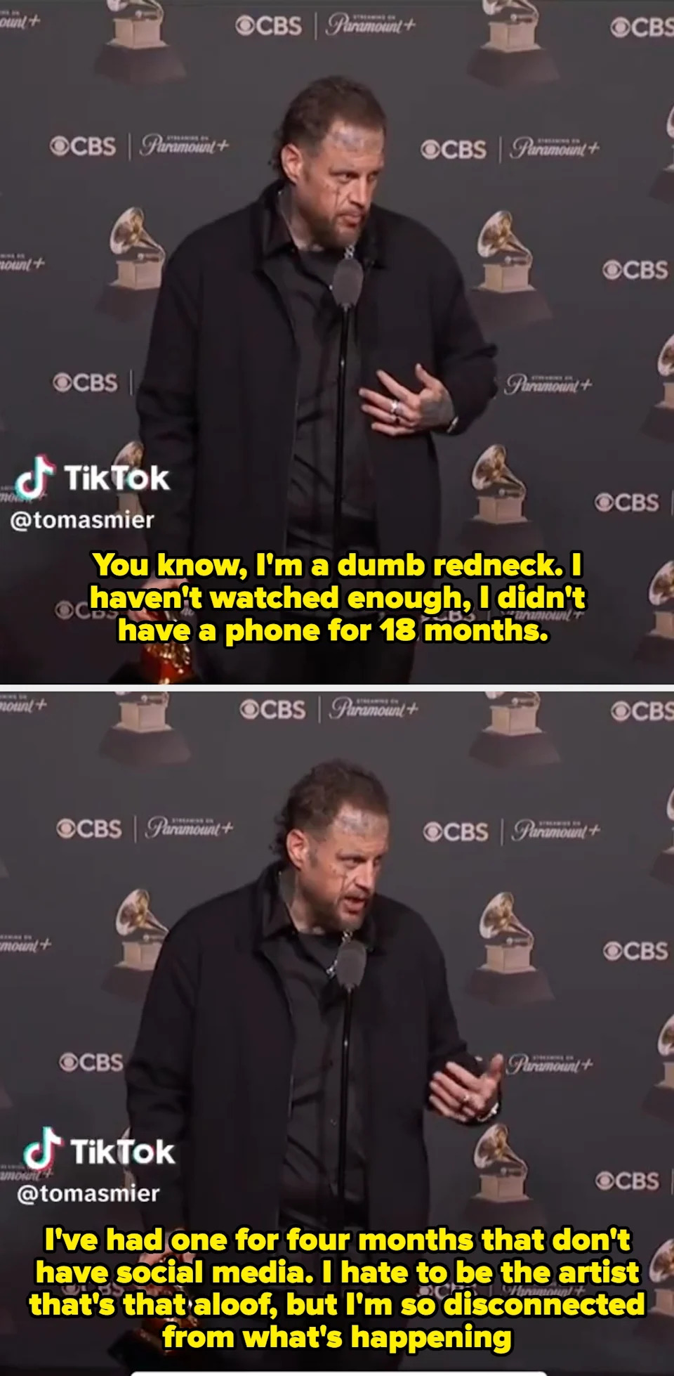 A man holds a Grammy award at a press event, speaking into a microphone. He wears a dark suit and shirt