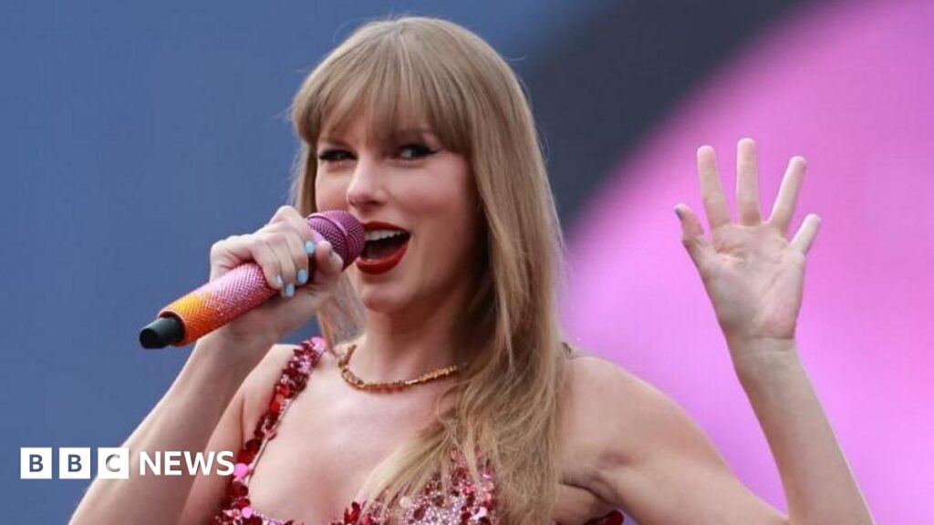 A close up shot of Taylor Swift singing on stage with a sparkly pink microphone while waving to the audience