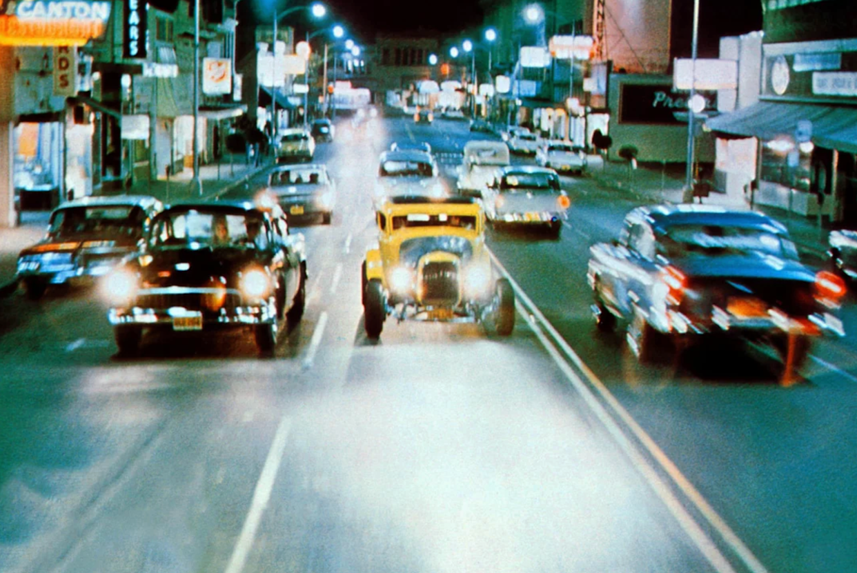 A bright yellow hot rod speeds down a lively city street at night, surrounded by classic cars under glowing streetlights and neon signs, creating a vibrant, retro atmosphere.