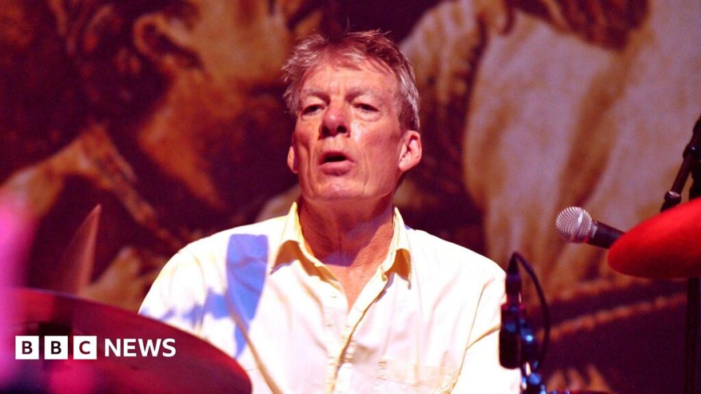 An older man with grey hair wearing a shirt and sitting behind a drum set and in front of a mural/picture.