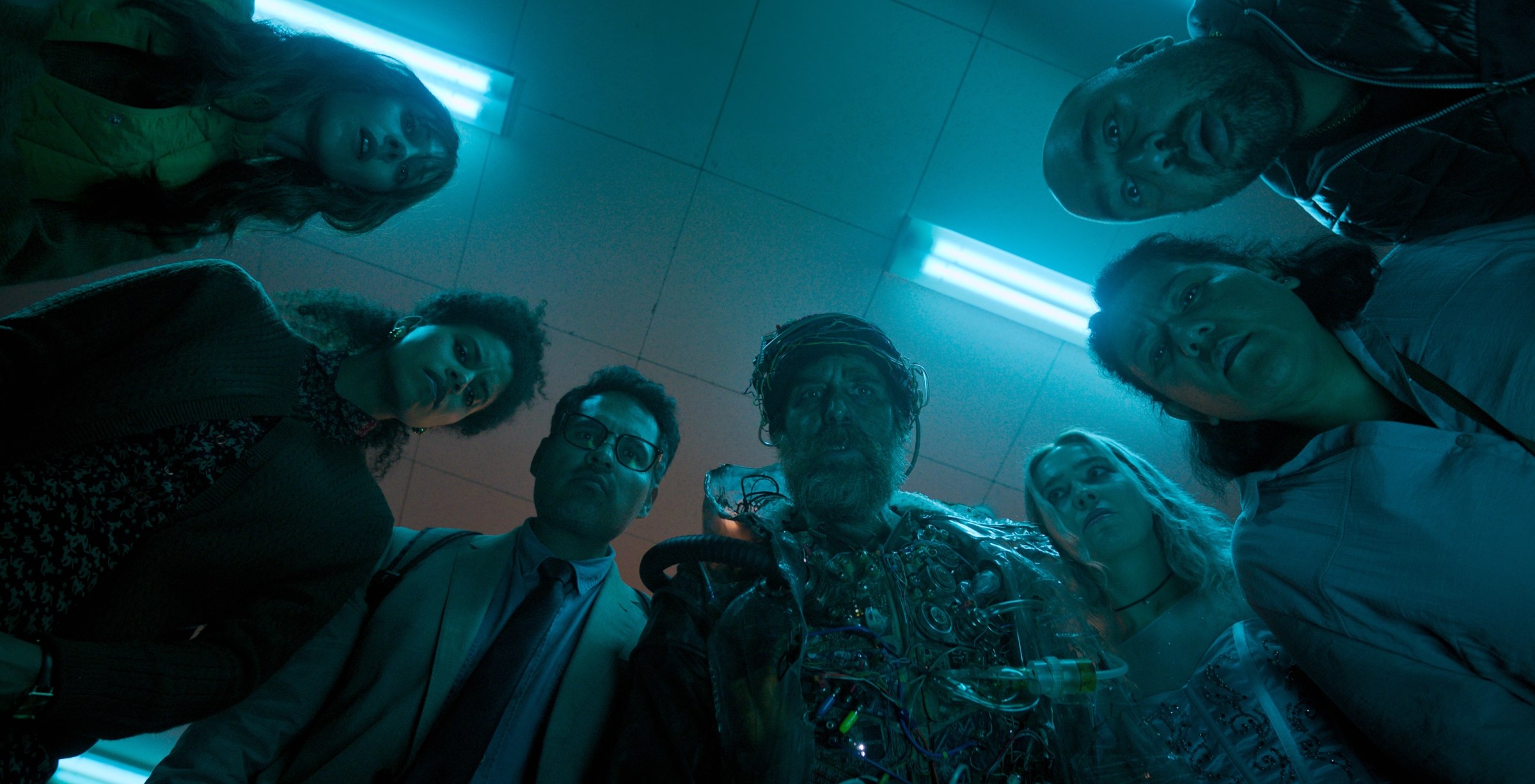 Four woman and three men standing together in a semicircle looking down at something on the floor in confusion.