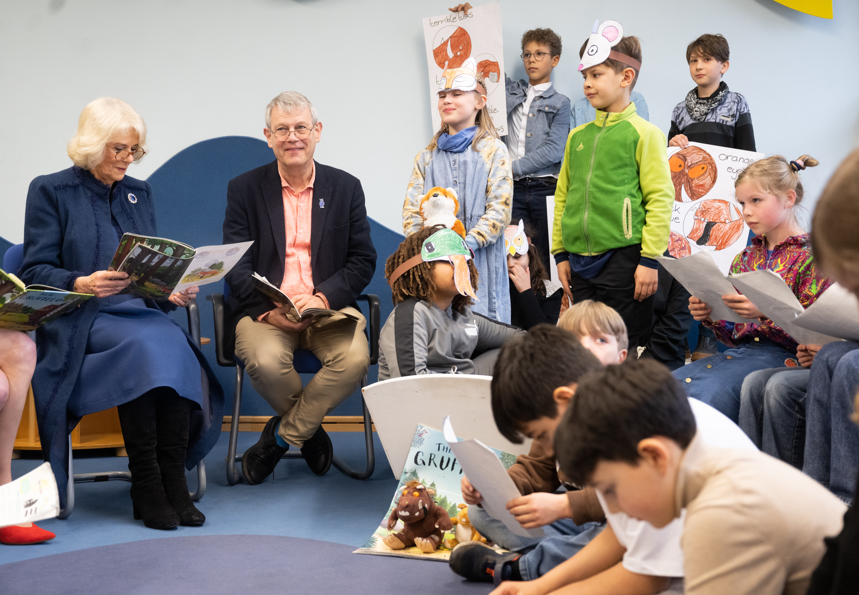 Queen Camilla reading a book sitting in a chair and surrounded by children