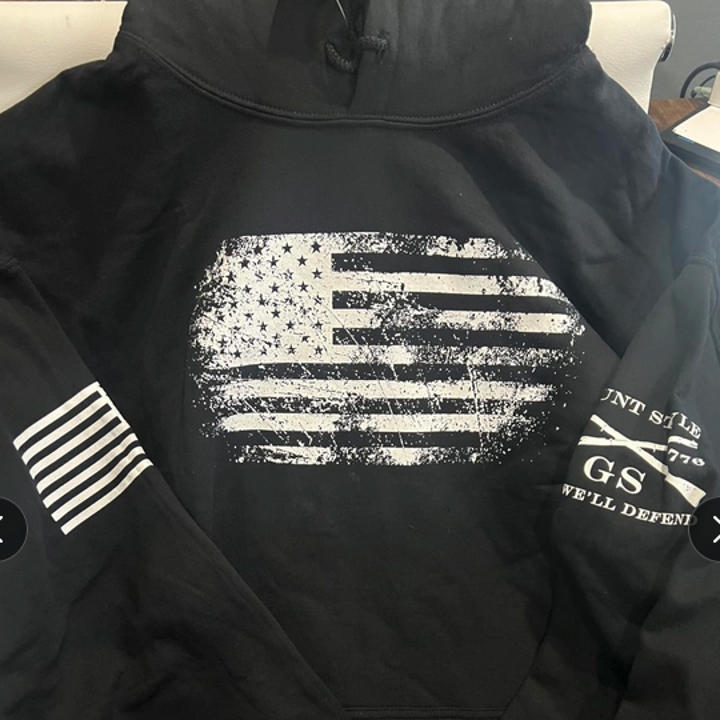 Black hoodie featuring an American flag design with grunge texture and text on the sleeves reading "GSTS" and "WE'LL DEFEND."