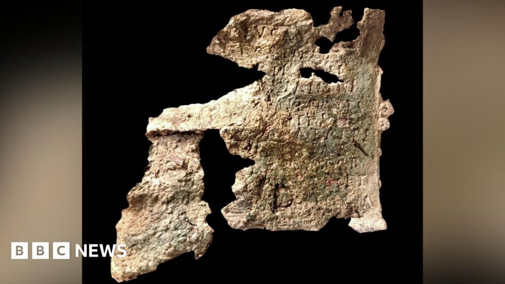 A damaged old bronze plaque on a dark background. Words etched in the metal can just be made out but the plaque has may holes in it.
