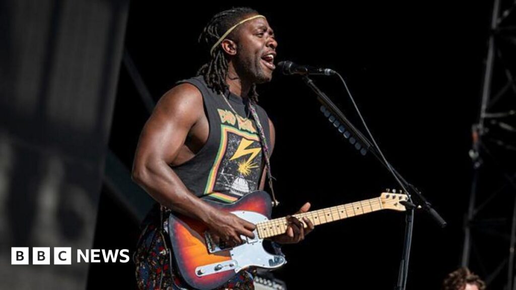 Muscly man in graphic tee vest top plays dark wood electric guitar on stage.