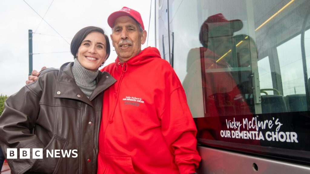 Vicky McClure and a man in a red 'Dementia Choir' jumper standing next to a tram with the words 'Vicky McClure's Our Dementia Choice' written on the side