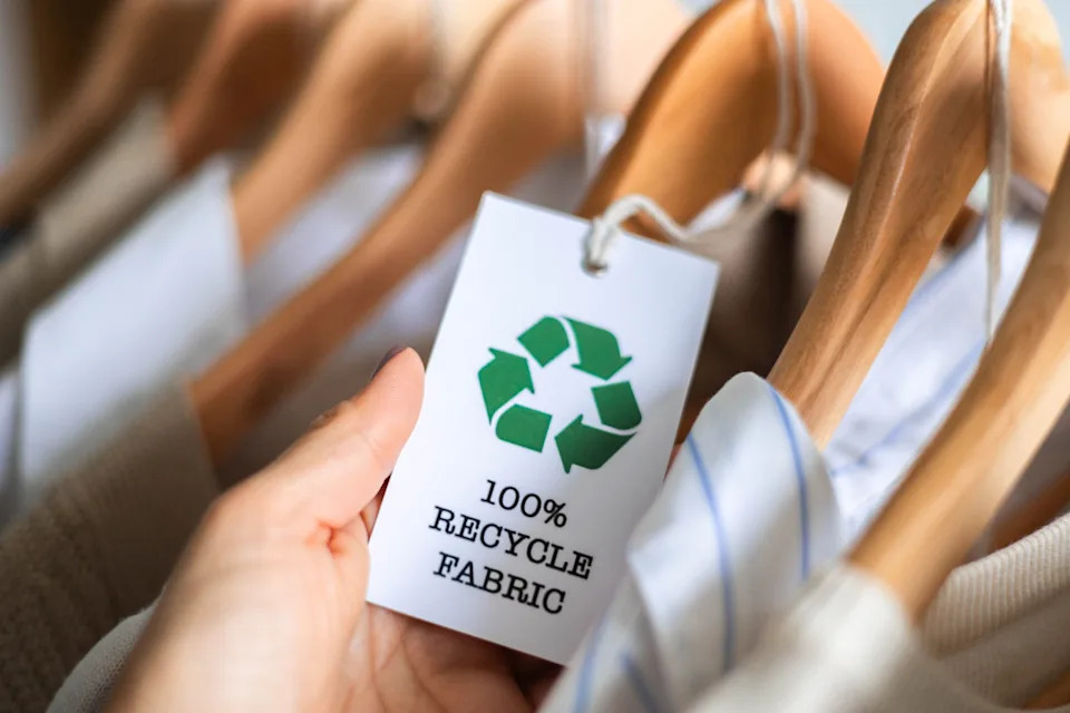Close-up of a recycled fabric clothing tag held in hand with sustainable clothes on wooden hangers. Eco-friendly fashion and ethical textile label for conscious consumers.