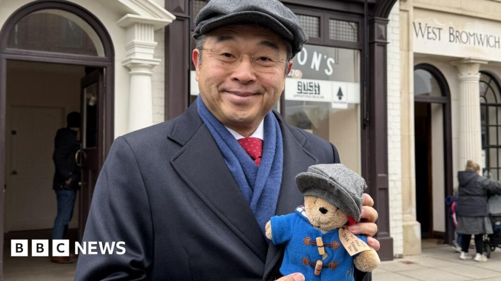 Hiroshi Suzuki has a grey flat cap on and a navy blue coat, with a blue scarf and red tie. He wears glasses and is holding a small Paddington Bear toy which also has a grey flat cap on, and a blue coat.