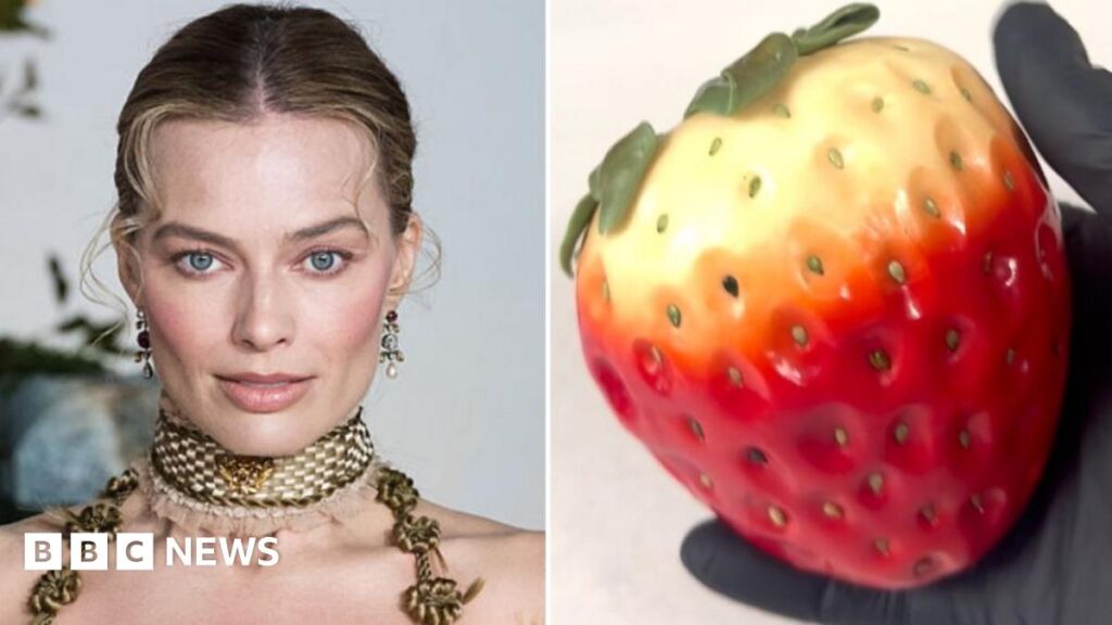 Margot Robbie has her blonde hair swept back with two curly tendrils at the front. She has dangly earrings and a gold choker-style thick necklace