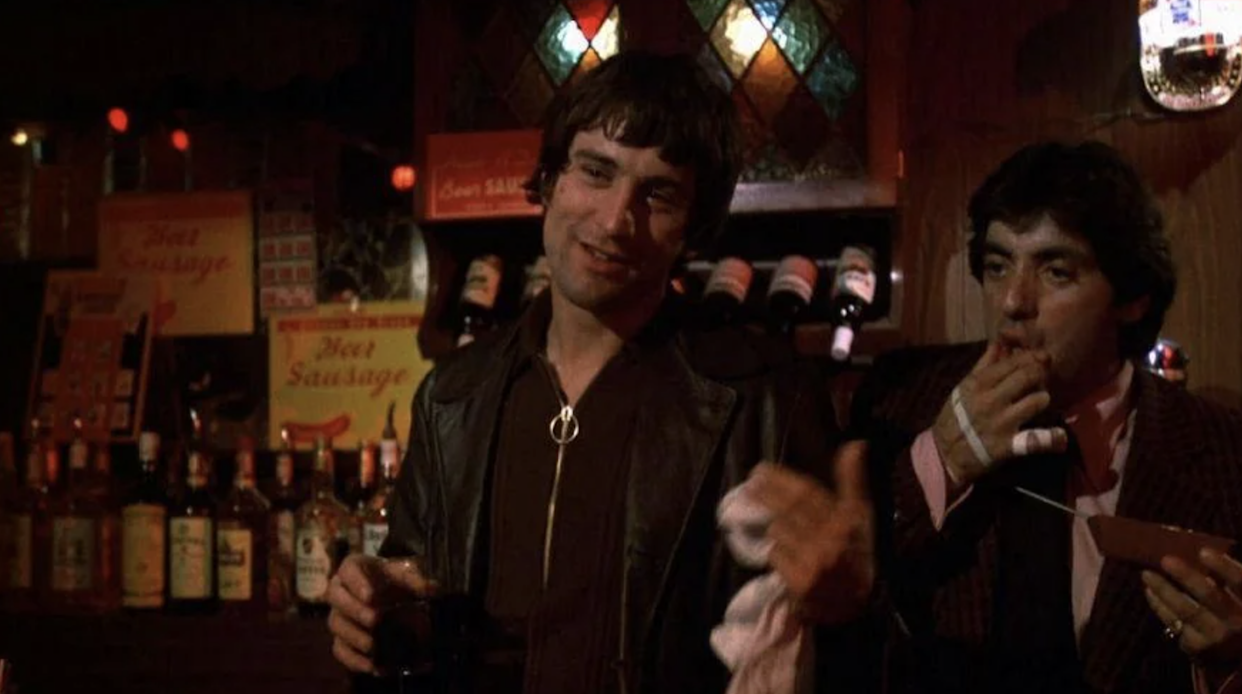 Two men stand at a dimly lit bar; one holds a drink and gestures with his hand, wearing a leather jacket and necklace, while the other eats food, holding a napkin, with bottles and stained glass in the background.