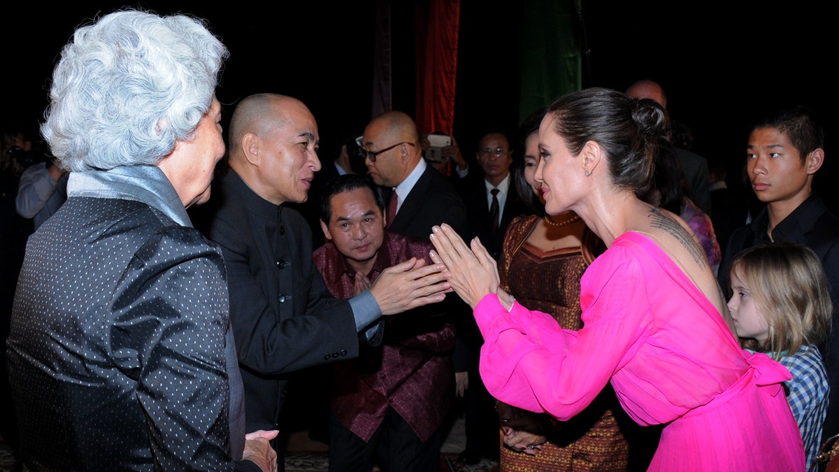 Angelina Jolie bows her head toward King Norodom Sihamoni while Queen Monique watches at an outdoor event.