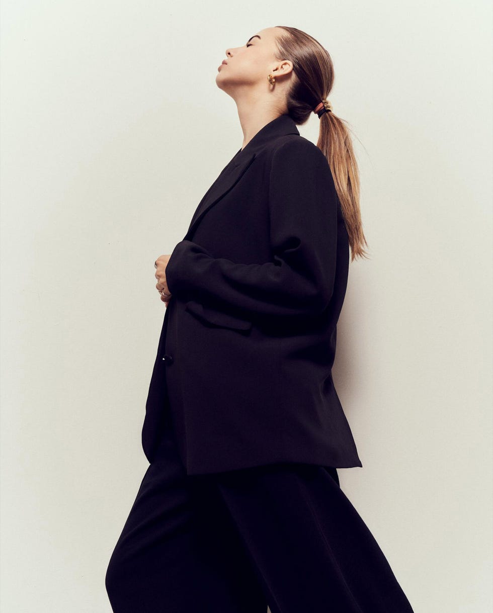 a figure in a tailored black outfit stands side profile against a light background a figure in a tailored black outfit stands side profile against a light background