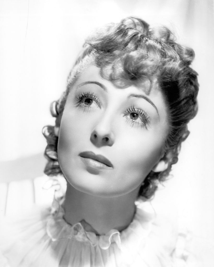 Luise Rainer looks up while wearing a high, frill-collared shirt or dress.