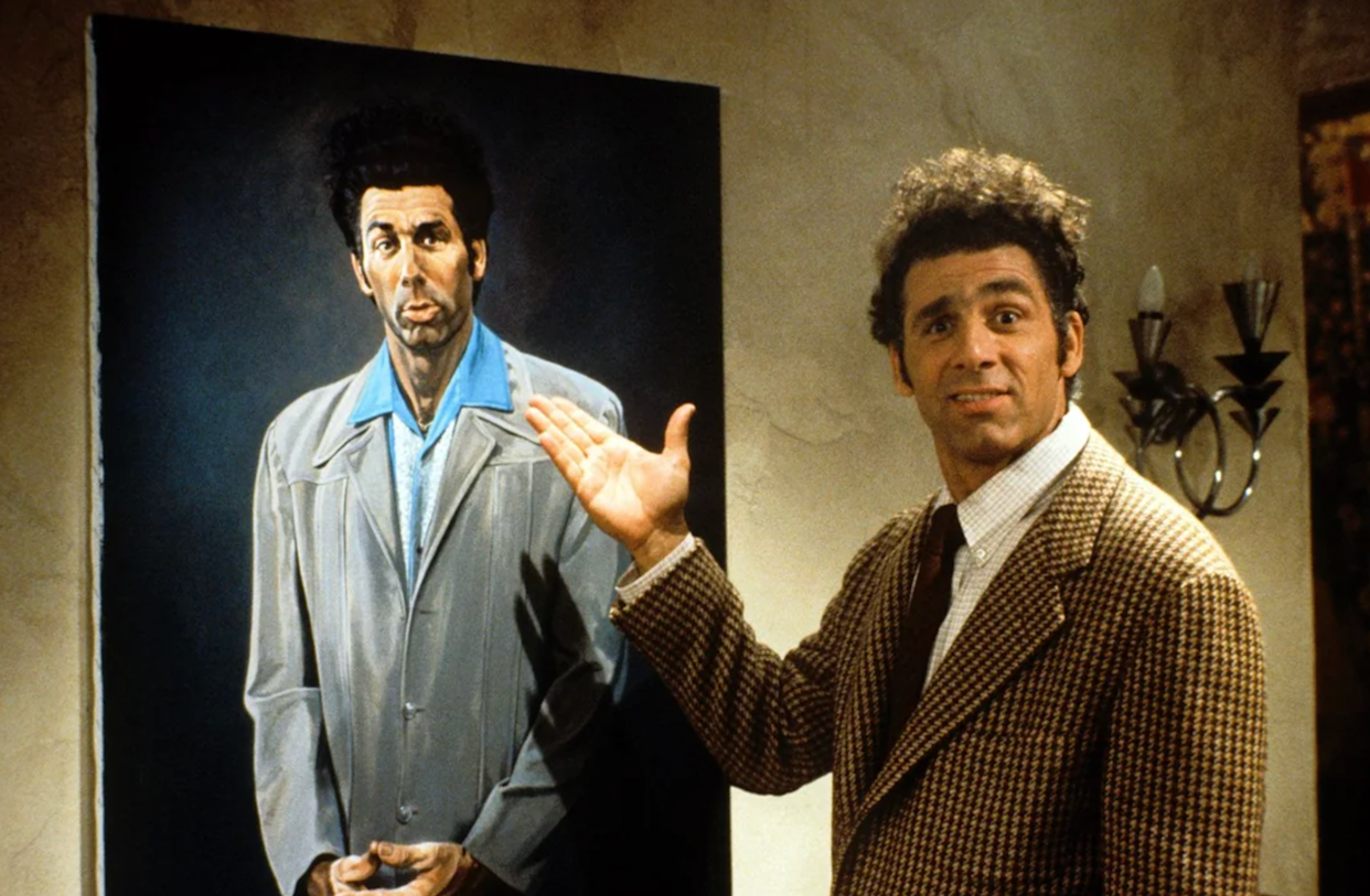 A man with curly hair and a patterned jacket stands next to a large painted portrait of himself in a blue shirt and gray coat, gesturing proudly toward the artwork on a beige wall.