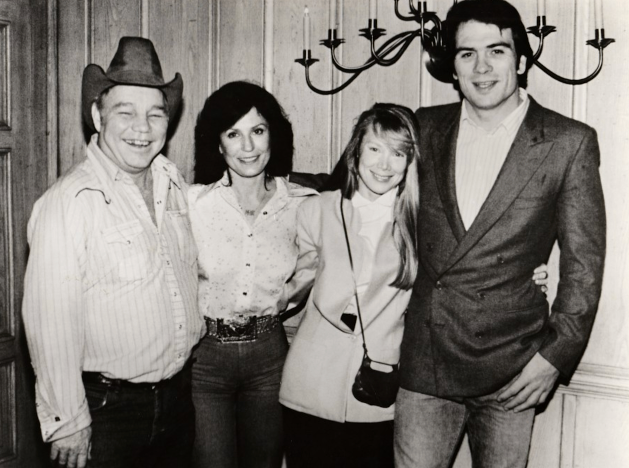 A black-and-white photo of two men and two women standing together and smiling indoors. One man wears a cowboy hat and Western shirt; the others wear jackets. A candle-style wall fixture is visible behind them.