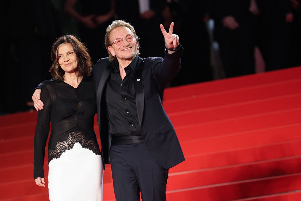 Ali Hewson and Bono stand on the red carpet together, Bono holds up a peace sign and has one arm around his wife's shoulders.