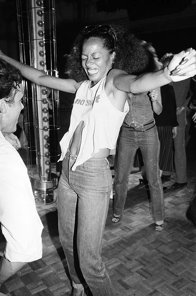 Person dancing energetically, wearing a sleeveless crop top and jeans, arms raised, in a lively setting