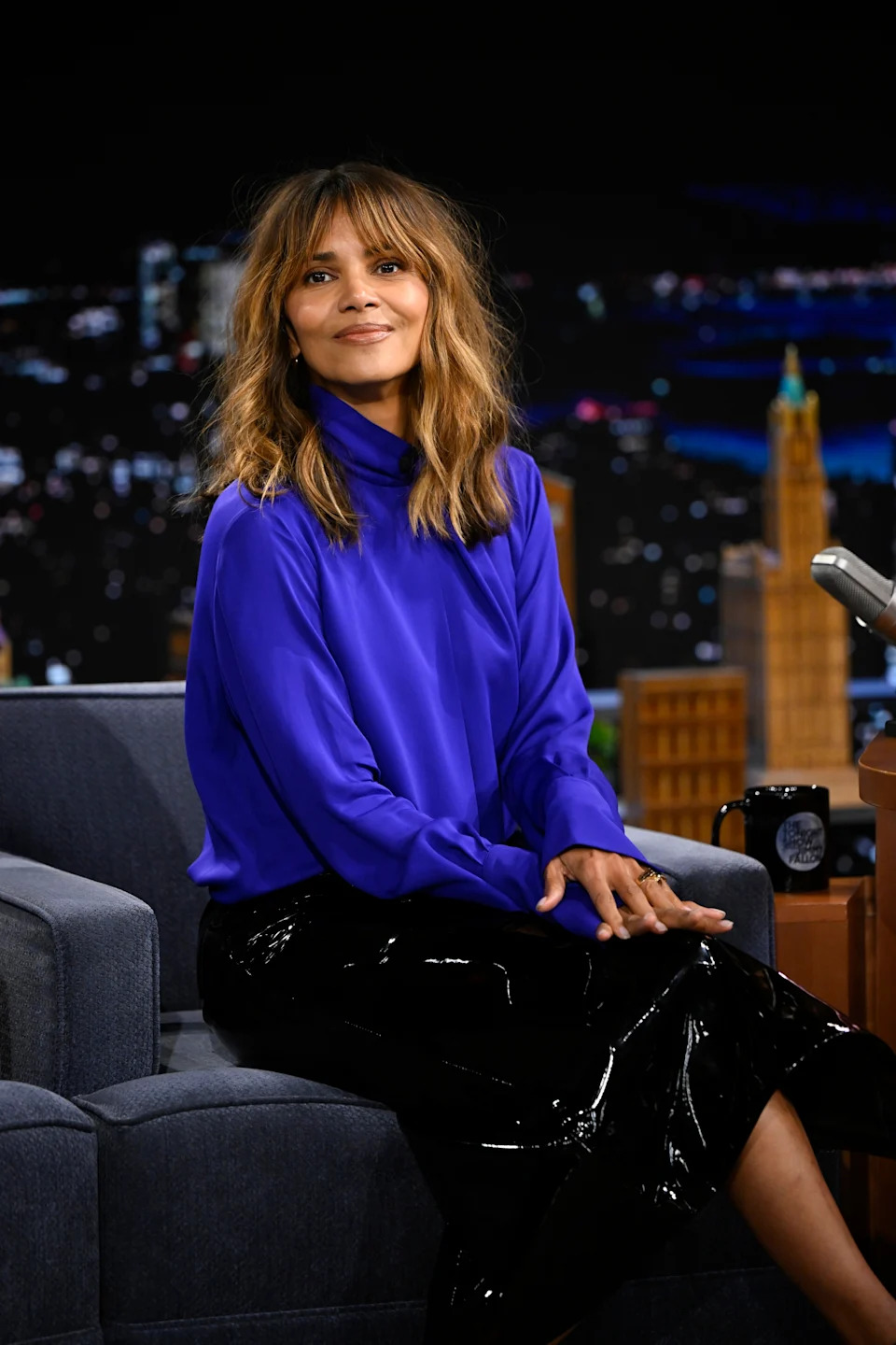 Halle Berry sits on The Tonight Show set wearing a glossy skirt and stylish long-sleeve top, with a cityscape backdrop visible